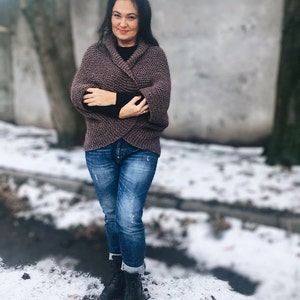 May include: A woman wearing a brown knitted cardigan with a shawl collar, blue jeans, and black boots. She is standing in front of a snowy background.