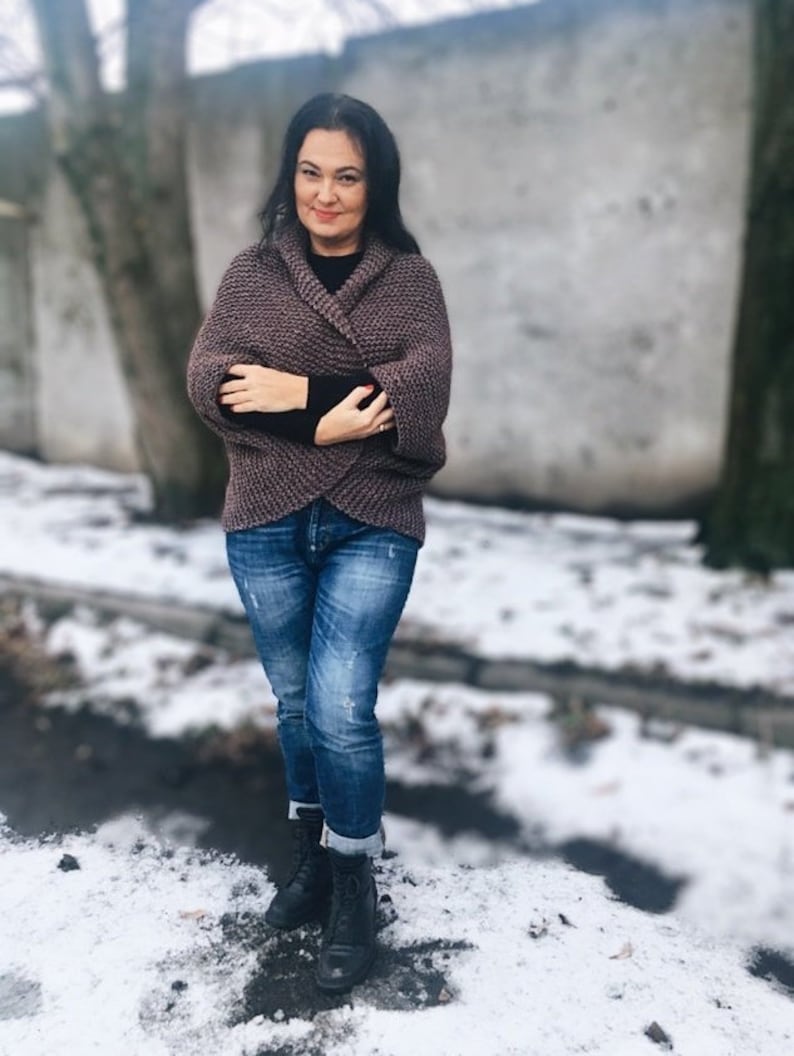 May include: A woman wearing a brown knitted cardigan with a shawl collar, blue jeans, and black boots. She is standing in front of a snowy background.