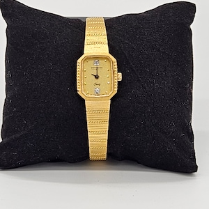 May include: A gold-tone Wittnauer watch with a square face and a diamond-encrusted bezel. The watch has a gold-tone band with a textured design.