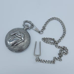 Puede incluir: Reloj de bolsillo plateado con cadena y cierre. La esfera del reloj presenta un diseño en relieve de una lengua y labios. La cadena está hecha de pequeños eslabones entrelazados.