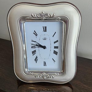 Może przedstawiać: Srebrny prostokątny zegar z białą tarczą i czarnymi rzymskimi cyframi. Zegar ma dekoracyjną srebrną ramę z ozdobnym górnym wzorem. Na tarczy zegara znajduje się napis "Florence".