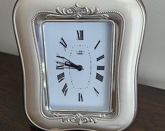 Può includere: Orologio rettangolare color argento con quadrante bianco e numeri romani neri. L'orologio ha una cornice decorativa argentata con un design superiore ornato. Sul quadrante c'è la scritta "Florence".