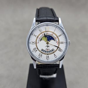 May include: A silver wristwatch with a white face, black leather strap, and Roman numeral hour markers. The watch features a moon phase indicator and the text "Луч" on the face. The watch is displayed on a black stand.