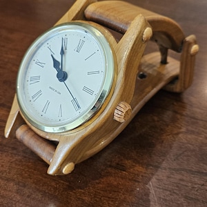 May include: A wooden clock with a gold-colored bezel and a cream-colored face. The clock has Roman numerals and black hands. The wooden frame is light brown and has a curved design. The clock is labeled "MADE IN USA".