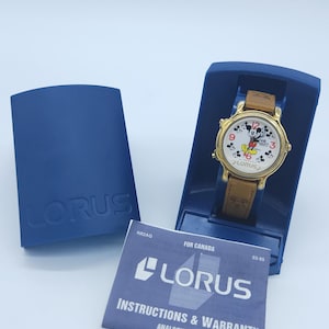 May include: A gold-tone Mickey Mouse watch with a brown leather strap. The watch face features a white background with Mickey Mouse in the centre. The watch is in a blue box with a Lorus instruction booklet.