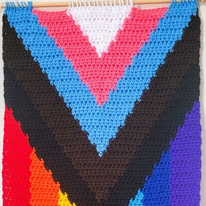 Puede incluir: Un tapiz de pared de crochet arcoíris con un patrón de chevron y flecos. Los colores son rojo, naranja, amarillo, verde, azul, índigo y violeta.