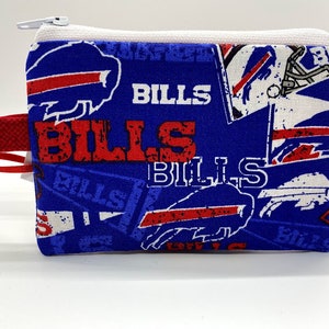 May include: A small, rectangular coin purse with a blue background and a repeating pattern of the word "BILLS" in red and white, along with the team logo. The purse has a white zipper and a red wrist strap with a key ring.