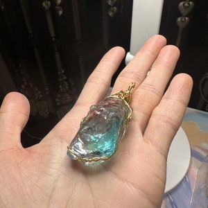May include: A blue and green crystal pendant wrapped in gold wire. The pendant is being held in a hand.