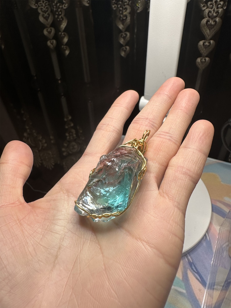 May include: A blue and green crystal pendant wrapped in gold wire. The pendant is being held in a hand.