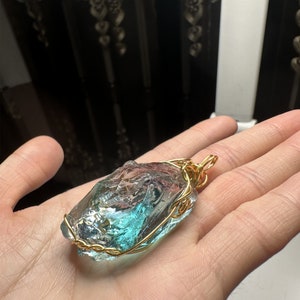 May include: A teal blue glass pendant wrapped in gold wire. The pendant is irregularly shaped and has a rough, natural texture.