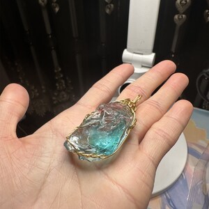 May include: A blue and green crystal pendant wrapped in gold wire. The pendant is being held in a hand.