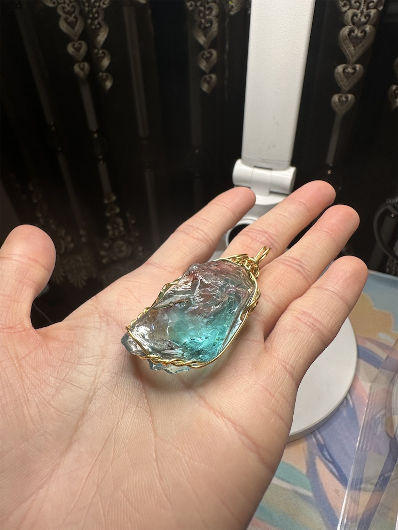 May include: A blue and green crystal pendant wrapped in gold wire. The pendant is being held in a hand.