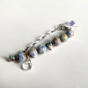 Op de afbeelding: Een armband met een zilverkleurige ketting verbonden aan een reeks ronde, gemarmerde blauwe en bruine kralen. Het ontwerp bevat een kleine zilveren hartvormige bedel, een doorzichtige halvemaanvormige hanger en een enkele paarse gefacetteerde kraal.