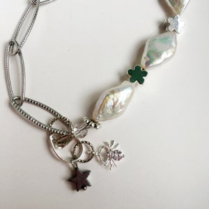 Op de afbeelding: Een zilveren ketting met een zilveren bedel met een ster, een spin en een kristallen kraal. De ketting heeft ook twee witte barokparels en twee groene bloemenbedels.