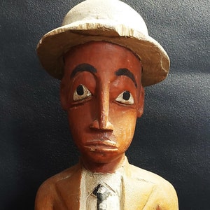 Könnte beinhalten: Eine Holzfigur eines Menschen, der einen braunen Anzug, ein weißes Hemd, eine schwarze Krawatte und einen weißen Hut trägt. Die Figur steht auf einem Holzsockel.