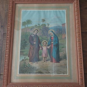 Puede incluir: Impresión religiosa enmarcada que representa una escena familiar con tres figuras con túnicas coloridas. La obra de arte presenta un detallado fondo de paisaje y está encerrada en un marco marrón ornamental. Una pieza vintage.