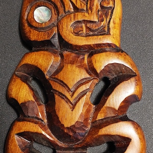 Könnte beinhalten: Eine braune Holz-Tiki-Schnitzerei mit einem Gesicht und zwei Augen aus irisierendem Perlmutt.