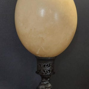 Peut inclure: Lampe décorative avec un grand abat-jour en forme d'œuf clair sur une base en métal sombre ornée. La base présente des volutes complexes et un design détaillé, pour une esthétique vintage. La hauteur de la lampe est d'environ 30 cm.