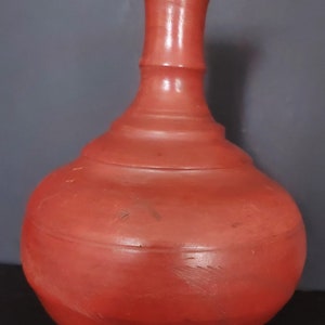 May include: A red clay vase with a wide base and a narrow neck. The vase is made of terracotta and has a rustic, handmade look.