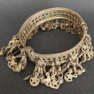 May include: Antique silver-toned hinged bracelet with intricate openwork and dangling charms. The bracelet features small bells and star-shaped charms, showcasing a vintage aesthetic. The metal has a slightly tarnished appearance, adding to its aged charm.