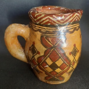 Peut inclure: Une petite cruche en céramique peinte à la main avec une anse. La cruche présente une base beige avec des motifs géométriques rouges et noirs. Le bord est orné d'un motif en zigzag. La anse est incurvée et la cruche a un petit bec.