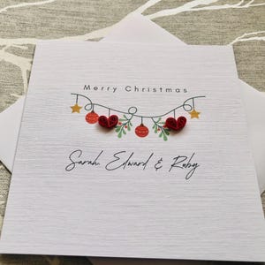 May include: A white Christmas card with a textured surface, featuring the words "Merry Christmas" above a garland of red ornaments, hearts, and stars. The names "Sarah, Edward & Ruby" are written below.