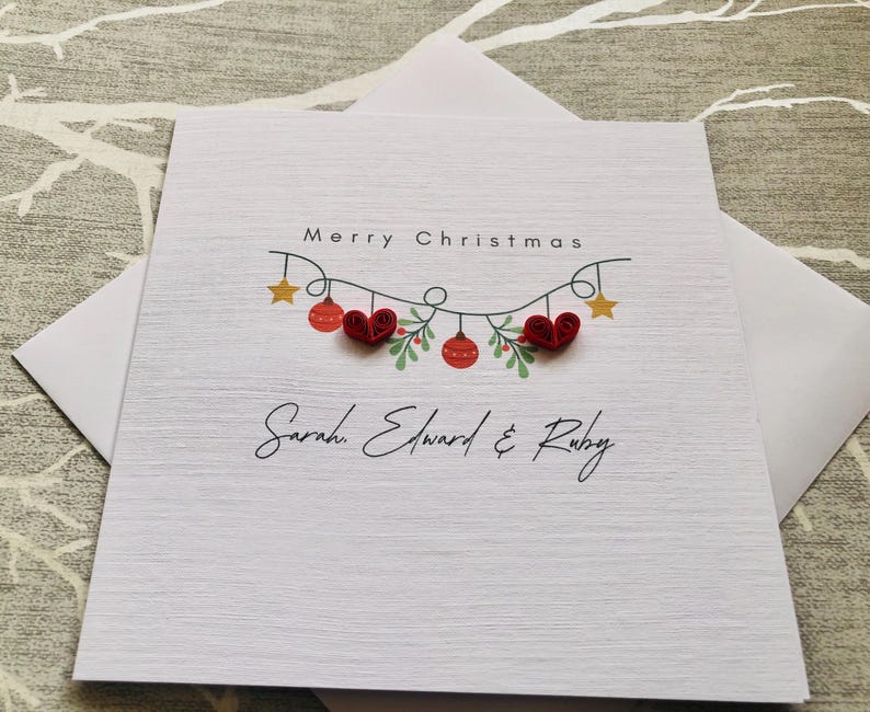 May include: A white Christmas card with a textured surface, featuring the words "Merry Christmas" above a garland of red ornaments, hearts, and stars. The names "Sarah, Edward & Ruby" are written below.