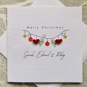 May include: A white textured Christmas card with the words "Merry Christmas" and the names "Sarah Edward & Ruby" in black script. The card features a garland of red ornaments, hearts, and green leaves.