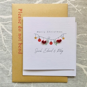 May include: A white Christmas card with the text "Merry Christmas" and the names "Sarah Edward & Ruby". The card features a garland of red ornaments, hearts, and stars. The card is on a brown envelope with the text "Please do not bend".