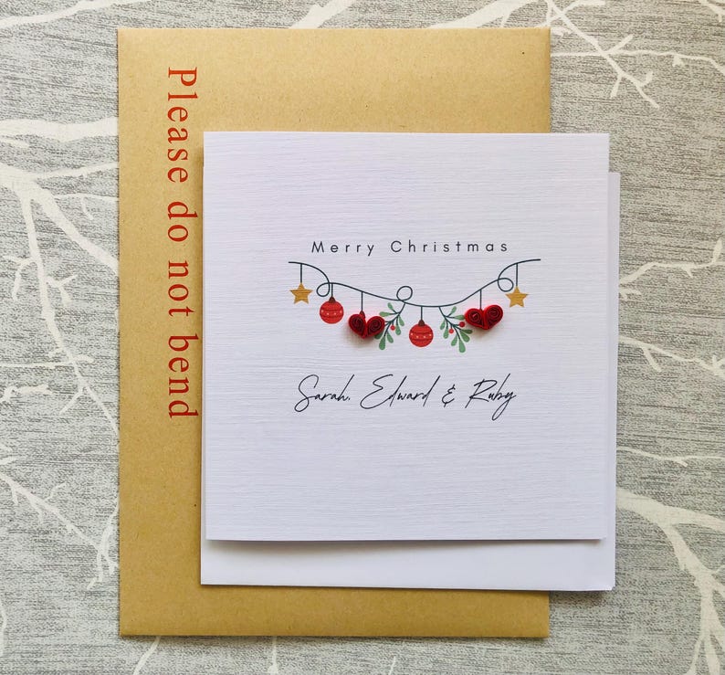 May include: A white Christmas card with the text "Merry Christmas" and the names "Sarah Edward & Ruby". The card features a garland of red ornaments, hearts, and stars. The card is on a brown envelope with the text "Please do not bend".