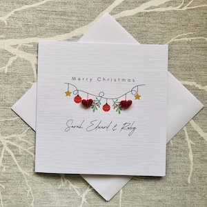 May include: A white Christmas card with the words "Merry Christmas" and the names "Sarah Edward & Ruby". The card features a garland of red hearts and ornaments, with gold stars and green leaves.