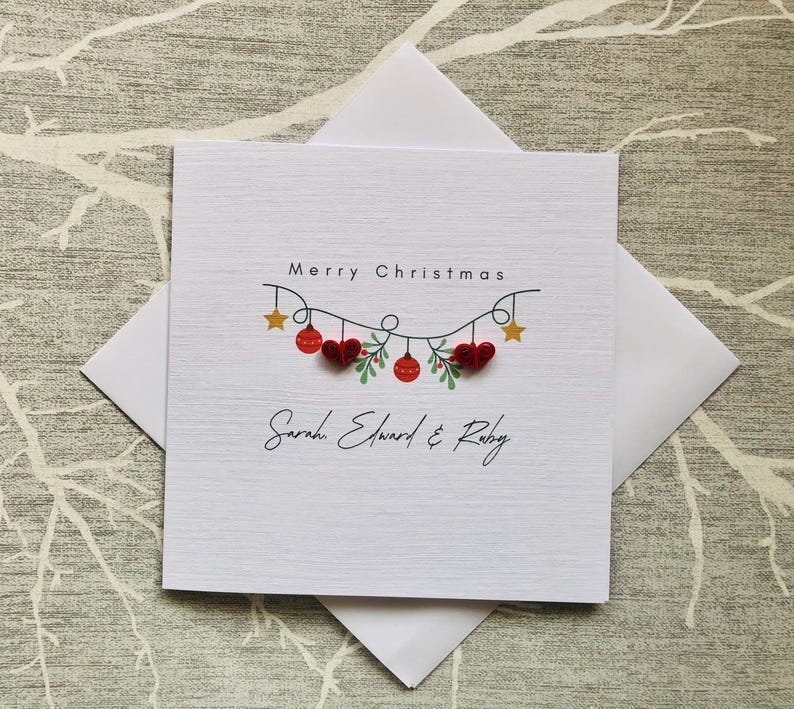 May include: A white Christmas card with the words "Merry Christmas" and the names "Sarah Edward & Ruby". The card features a garland of red hearts and ornaments, with gold stars and green leaves.