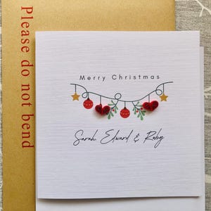May include: A white Christmas card with the words "Merry Christmas" and a garland of red ornaments, hearts, and stars. The names "Sarah, Edward & Ruby" are written below the garland. The card is in a brown envelope that says "Please do not bend".