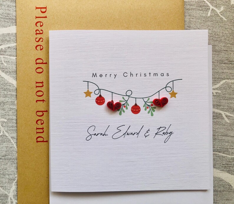 May include: A white Christmas card with the words "Merry Christmas" and a garland of red ornaments, hearts, and stars. The names "Sarah, Edward & Ruby" are written below the garland. The card is in a brown envelope that says "Please do not bend".