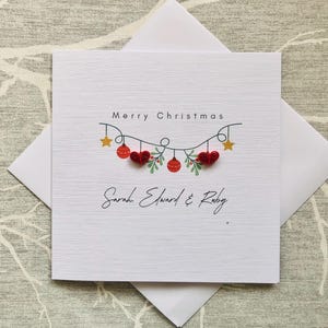 May include: A white Christmas card with a textured surface, featuring the text "Merry Christmas" above a garland of red hearts, ornaments, and green sprigs. The names "Sarah Edward & Ruby" are written below. The card is inside a white envelope.