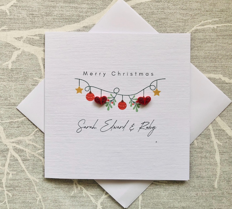 May include: A white Christmas card with a textured surface, featuring the text "Merry Christmas" above a garland of red hearts, ornaments, and green sprigs. The names "Sarah Edward & Ruby" are written below. The card is inside a white envelope.