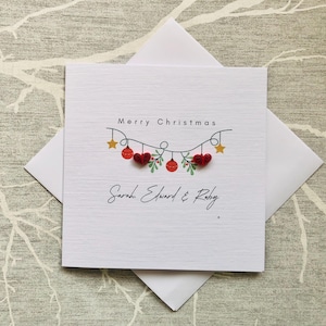 May include: White Christmas card with the words "Merry Christmas" and a decorative garland of red hearts, ornaments, and stars. The names "Sarah Edward & Ruby" are written below the garland. The card is inside a white envelope.