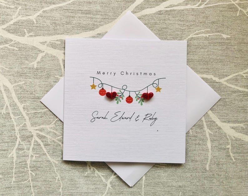 May include: White Christmas card with the words "Merry Christmas" and a decorative garland of red hearts, ornaments, and stars. The names "Sarah Edward & Ruby" are written below the garland. The card is inside a white envelope.