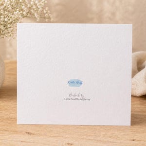 May include: A white square card with the text "Little Stuff" in a light blue watercolor font. The card is on a wooden surface, with a vase of white flowers to the left and a beige cloth to the right.