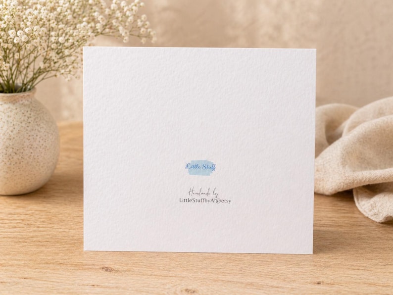 May include: A white square card with the text "Little Stuff" in a light blue watercolor font. The card is on a wooden surface, with a vase of white flowers to the left and a beige cloth to the right.
