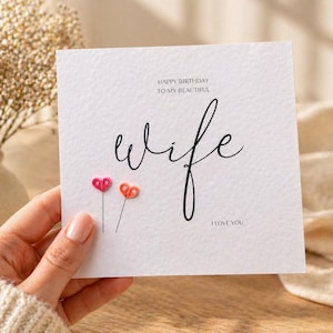 May include: A white greeting card with the words "Happy Birthday to my beautiful wife" and "I love you" in black script. Two small, colorful heart-shaped embellishments are attached to the card. A vase of dried flowers is in the background.