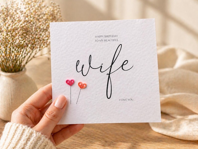 May include: A white greeting card with the words "Happy Birthday to my beautiful wife" and "I love you" in black script. Two small, colorful heart-shaped embellishments are attached to the card. A vase of dried flowers is in the background.
