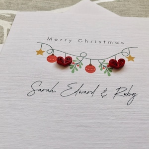 May include: A white Christmas card with the words "Merry Christmas" and a garland of ornaments and stars. The card features red heart-shaped embellishments and the names "Sarah, Edward & Ruby" in elegant script.
