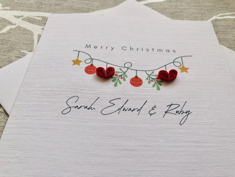 May include: A white Christmas card with the words "Merry Christmas" and a garland of ornaments and stars. The card features red heart-shaped embellishments and the names "Sarah, Edward & Ruby" in elegant script.