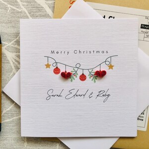 May include: A white Christmas card with the text "Merry Christmas" and the names "Sarah, Edward & Ruby" in script. The card features a garland of red hearts, ornaments, and gold stars. A brown pen and white envelope are next to the card.