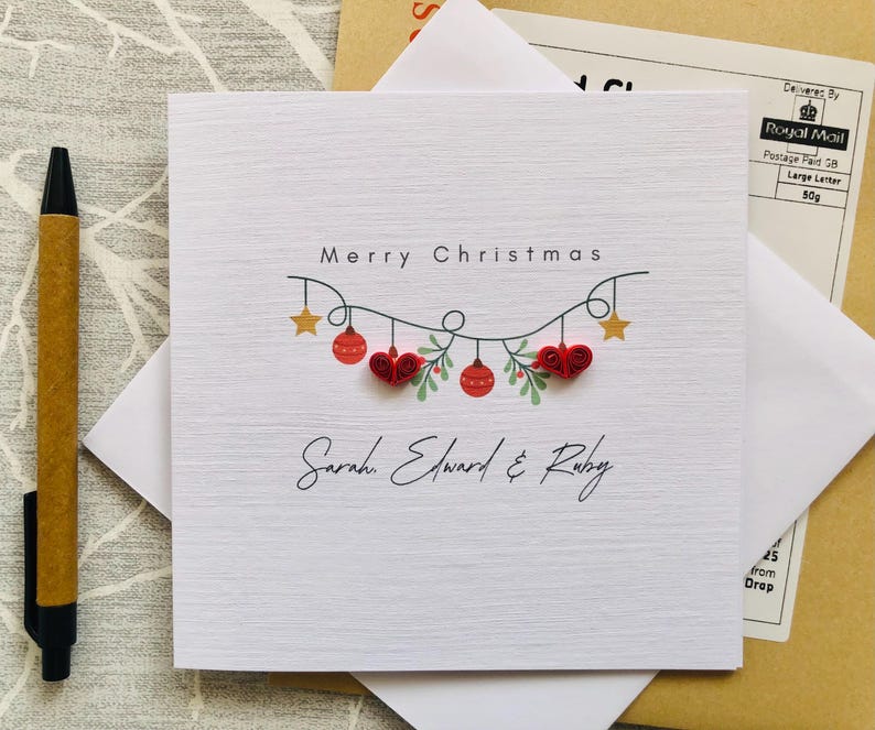 May include: A white Christmas card with the text "Merry Christmas" and the names "Sarah, Edward & Ruby" in script. The card features a garland of red hearts, ornaments, and gold stars. A brown pen and white envelope are next to the card.