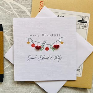 May include: A square white Christmas card with the words "Merry Christmas" and the names "Sarah, Edward & Ruby". The card features a garland of red ornaments and gold stars. A pen and envelope are also visible.