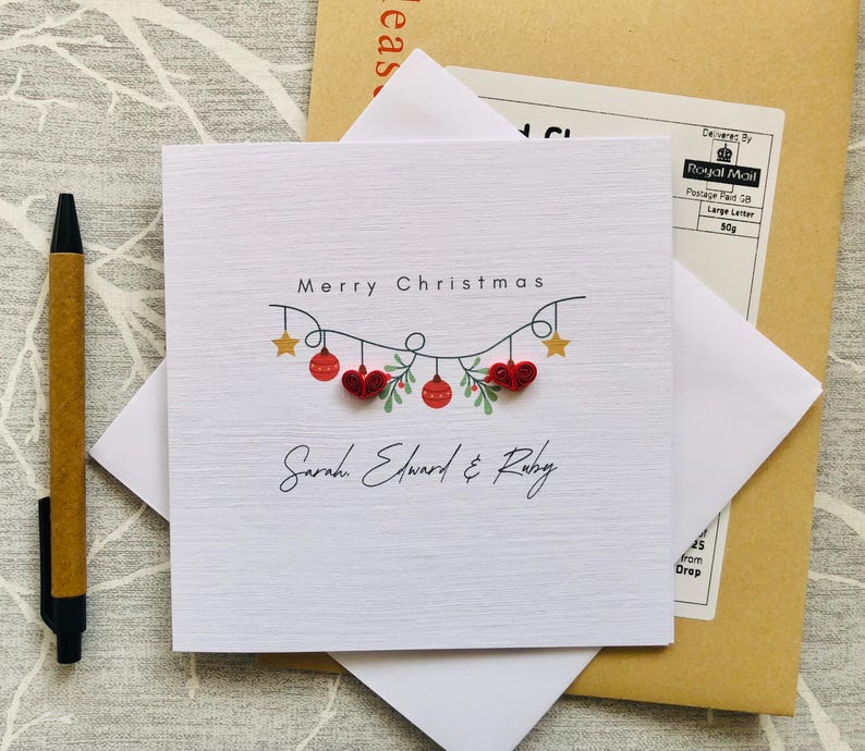 May include: A square white Christmas card with the words "Merry Christmas" and the names "Sarah, Edward & Ruby". The card features a garland of red ornaments and gold stars. A pen and envelope are also visible.