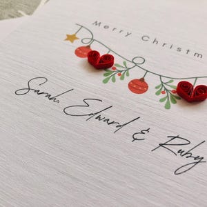 May include: A white Christmas card with the words "Merry Christmas" and the names "Sarah, Edward & Ruby" in elegant script. The card features a garland with red ornaments, green leaves, and two red heart-shaped embellishments.
