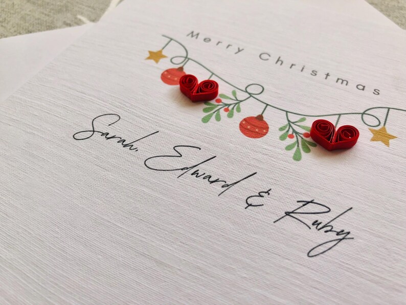 May include: A white Christmas card with the words "Merry Christmas" and the names "Sarah, Edward & Ruby" in elegant script. The card features a garland with red ornaments, green leaves, and two red heart-shaped embellishments.